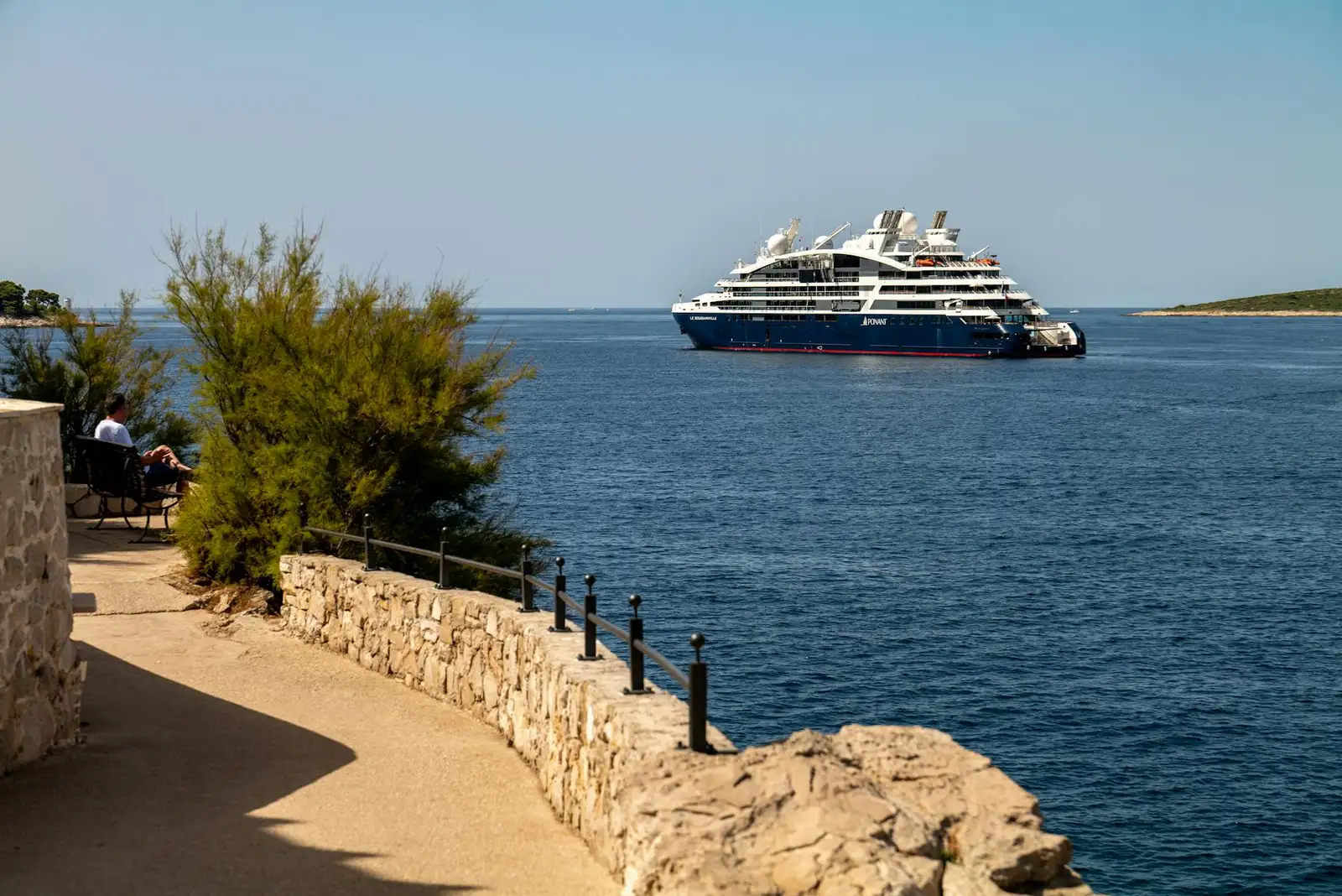 A luxury cruise ship sails near Primošten's scenic rocky coastline under a clear blue sky.